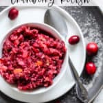 Cranberry relish in small white dish on small white plate on larger gray plate, with silver spoon and a few whole cranberries on the side