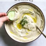 Lemon dill hummus in beige serving dish with olive oil drizzled on top, with golden spoon, and hand dipping a cucumber slice into the hummus