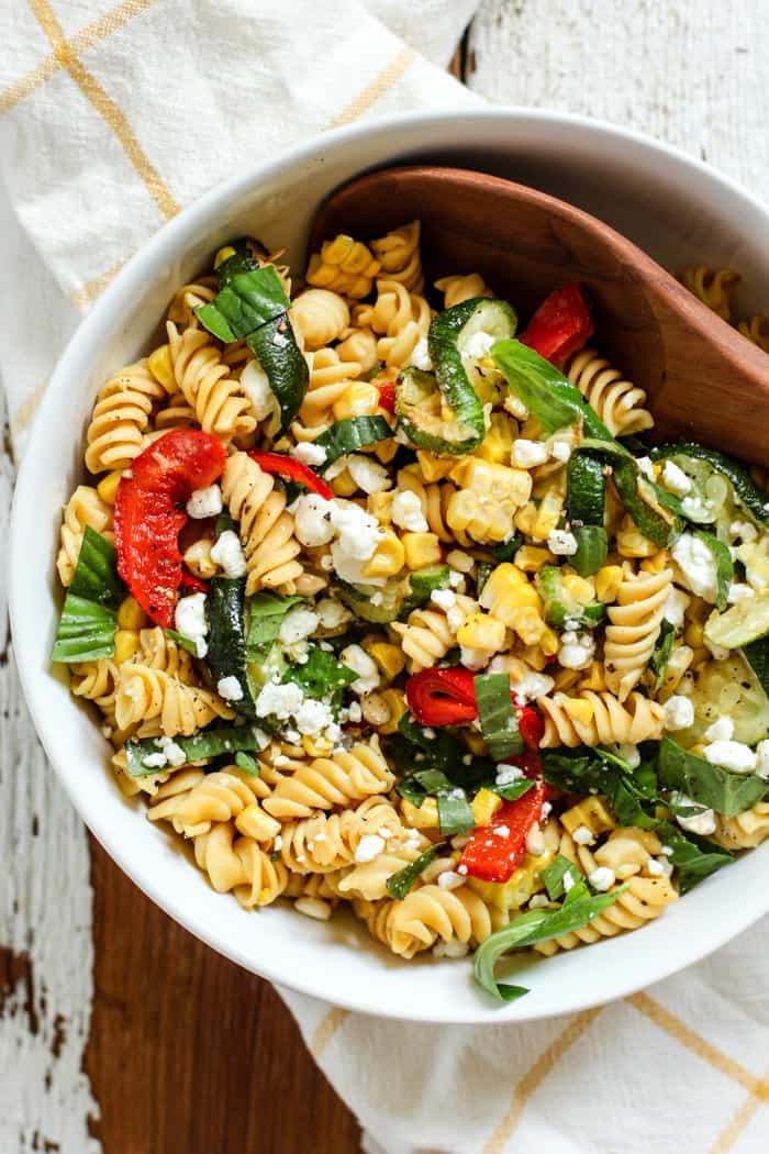 Late summer veggie pasta salad with red peppers, corn, zucchini, fresh basil, and goat cheese, in a white bowl with a wooden spoon