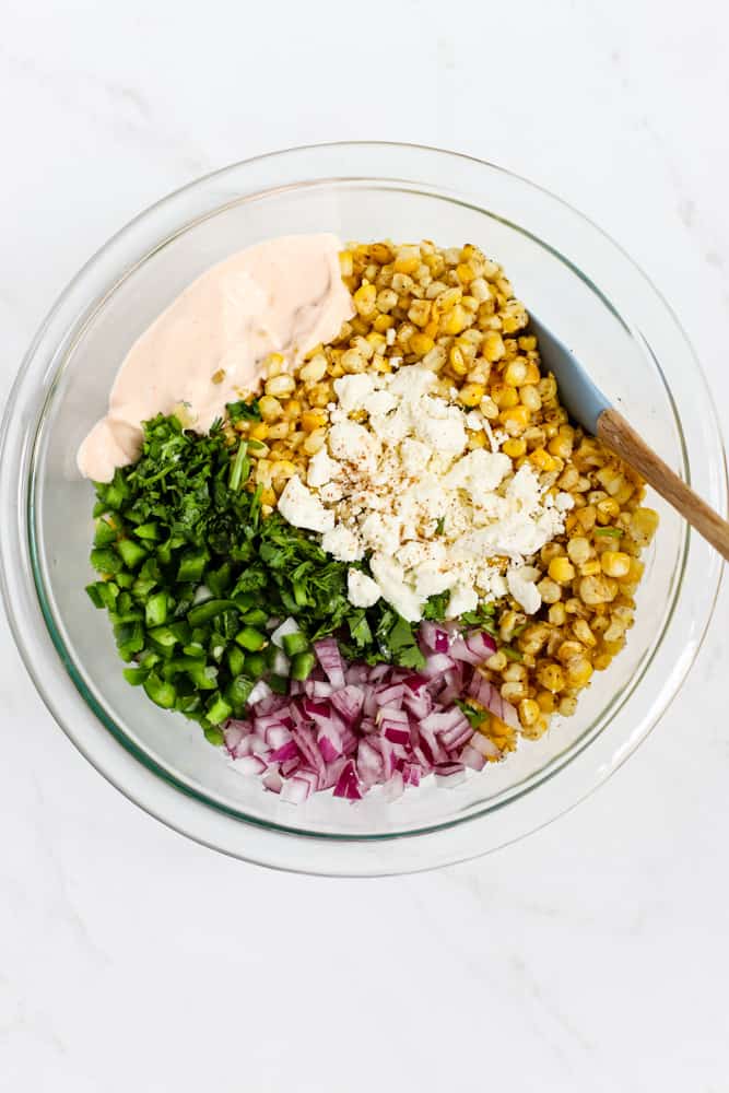 Roasted corn kernels, feta, chipotle seasoning, Greek yogurt, jalapeños, cilantro, and red onion in a glass bowl