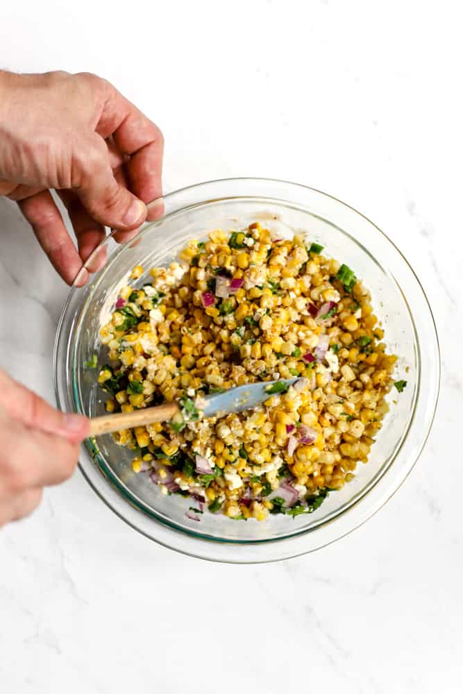 Hands mixing spicy Mexican street corn salad in a glass bowl using a small spatula
