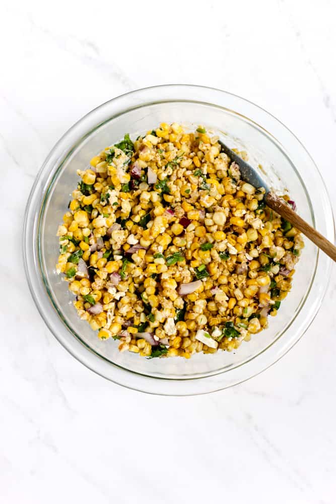 Spicy Mexican street corn salad mixed in a glass bowl with small spatula on white surface