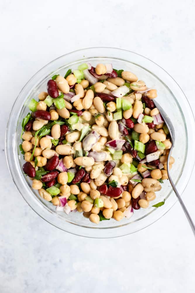 Modern three bean salad ingredients all in a clear glass bowl, tossed in creamy apple cider vinaigrette just poured in center, prior to tossing salad
