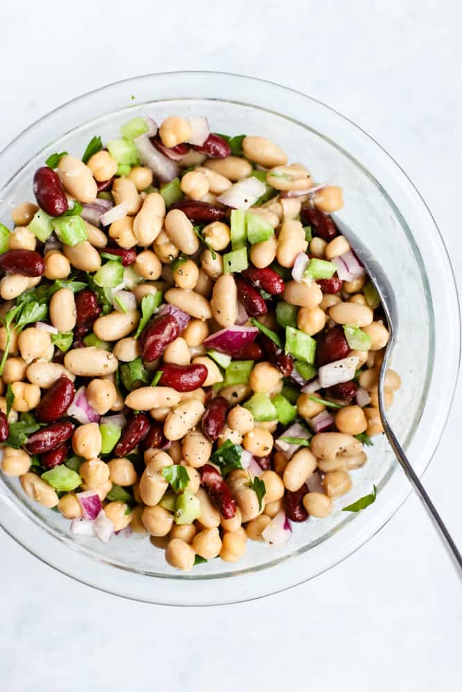 Modern three bean salad ingredients all in a clear glass bowl, tossed in creamy apple cider vinaigrette and topped with freshly cracked black pepper