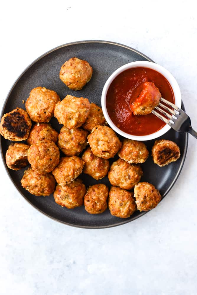 Baked Spanish chicken meatballs on dark gray plate with small white bowl of pizza sauce, one meatball on fork dipped in sauce