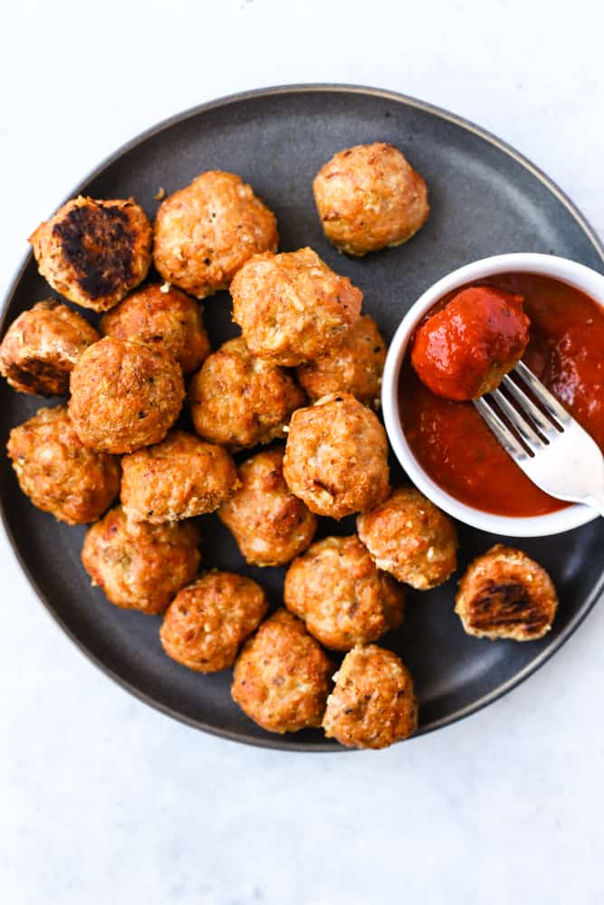 Baked Spanish chicken meatballs on dark gray plate with small white bowl of pizza sauce, one meatball on fork dipped in sauce