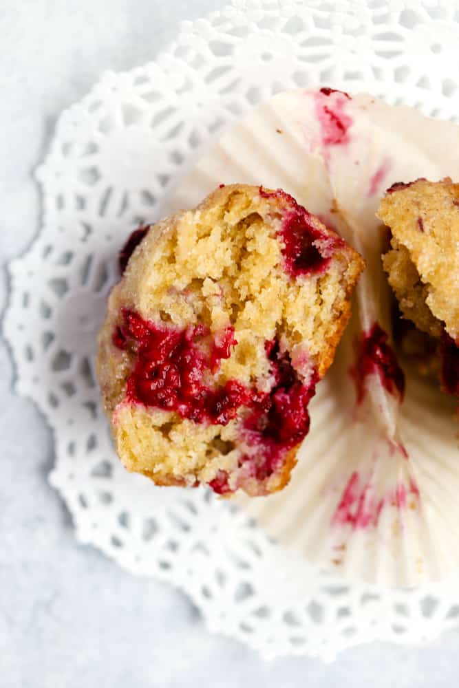 One wholesome raspberry lemon muffin cut open and left in parchment muffin paper on white doily, with tender inner crumb and juicy raspberries showing