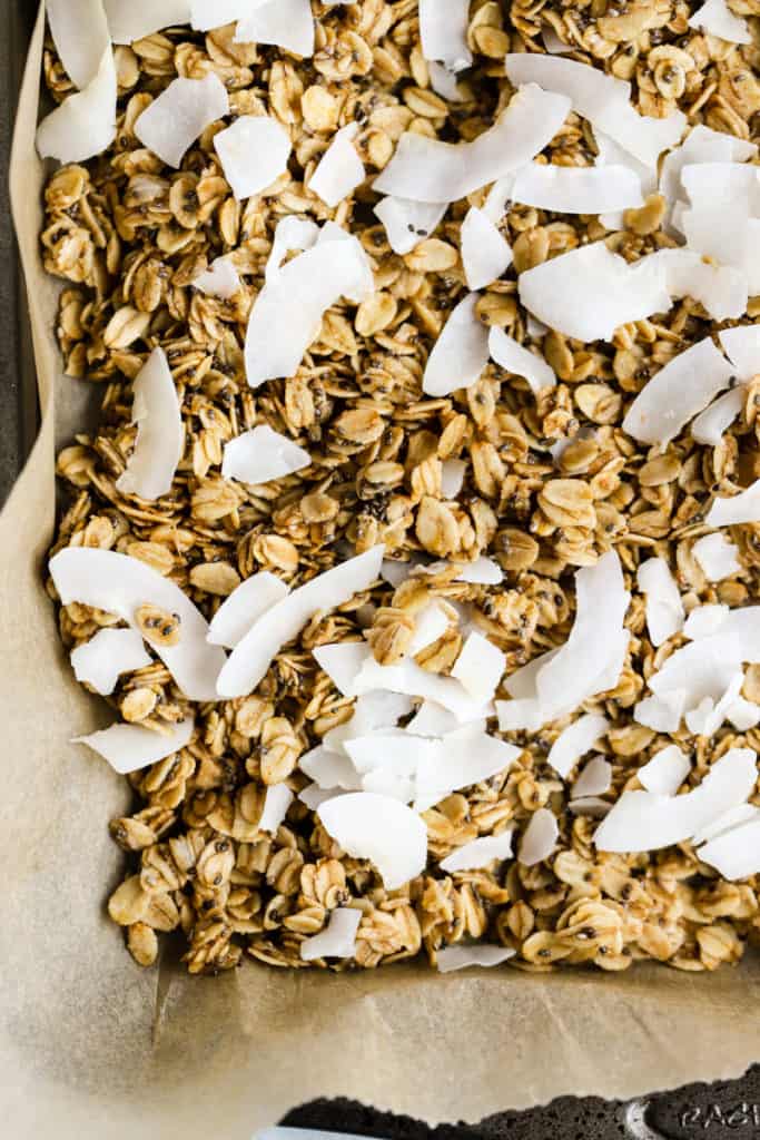 Simple coconut chia granola on parchment paper lined sheet pan with coconut just added before putting back in oven to toast