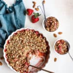 Healthy strawberry crisp in cream-colored pie dish with copper serving spoon and two small white bowls with more crisp, with almonds and fresh strawberries on the side