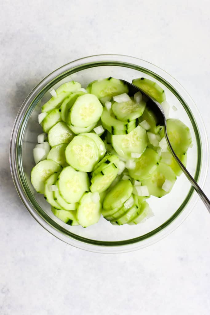Cucumber slices with a few pieces of yellow onion diced small in clear glass bowl with spoon