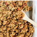 Healthy pumpkin granola in parchment paper-lined sheet pan with wooden spoon stirring the granola cluster