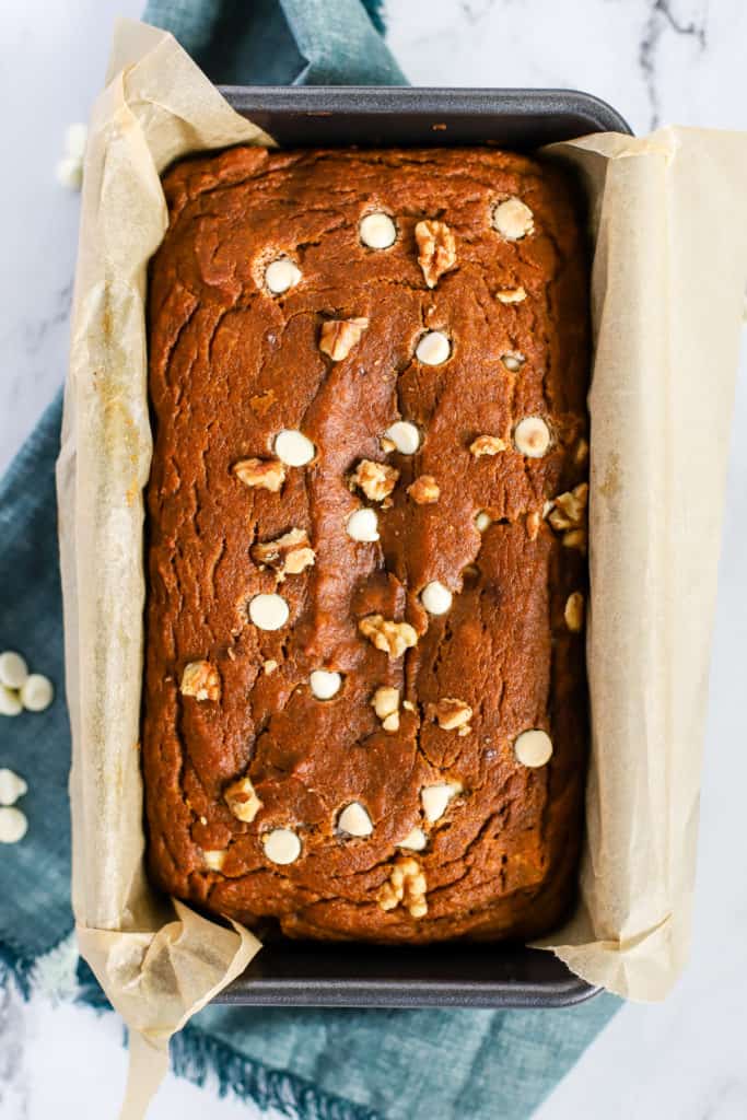 Fully baked healthy spelt pumpkin bread in bread pan lined with parchment paper, on teal linen on white marble surface