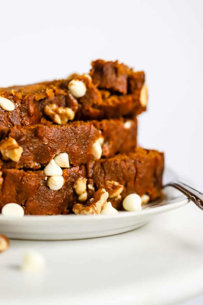 Stack of healthy spelt pumpkin bread slices on a small white plate with fork, on white marble surface, with a few walnuts and white chocolate chips sprinkled on the side