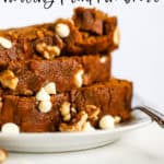Stack of healthy spelt pumpkin bread slices on a small white plate with fork, on white marble surface, with a few walnuts and white chocolate chips sprinkled on the side
