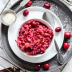 Cranberry relish in small white dish on small white plate on larger gray plate, with silver spoon and a few whole cranberries on the side