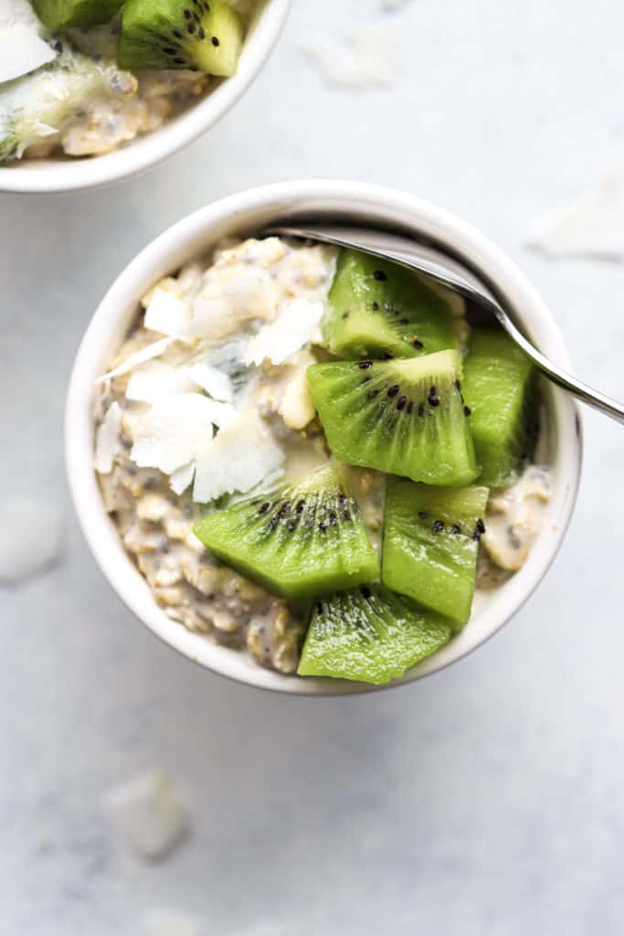 Overnight oats in small white bowl with spoon, topped with coconut flakes and diced kiwi, on white and light blue background