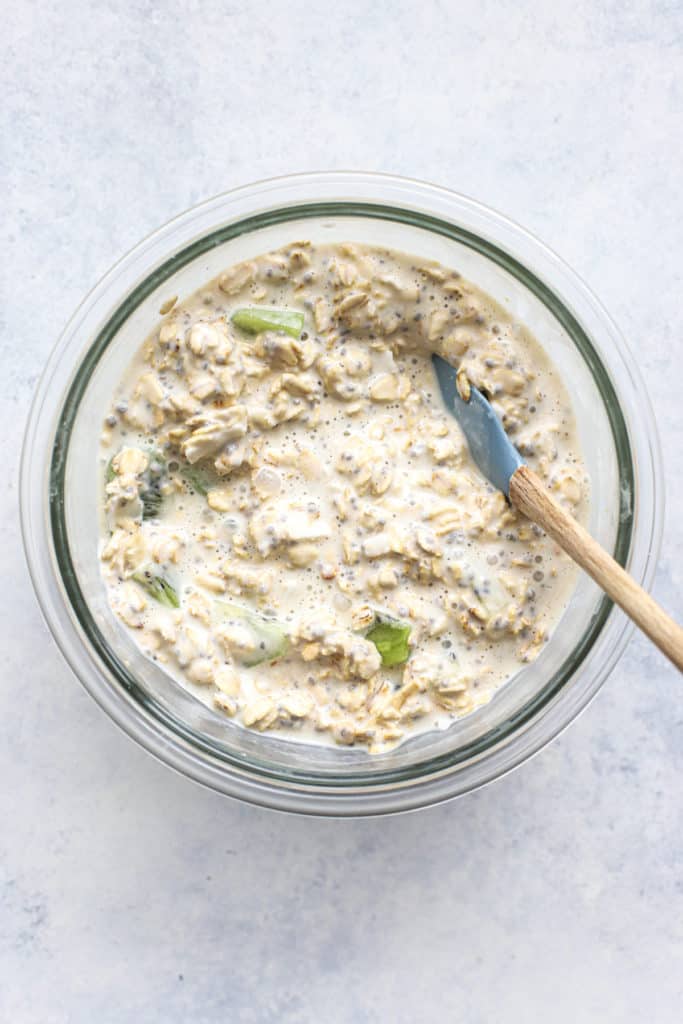Kiwi coconut overnight oats mixed together in clear glass bowl with small rubber spatula, on light blue and white surface