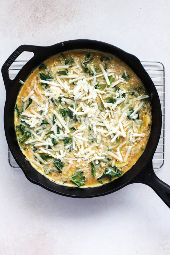 Egg mixture poured into black cast iron skillet with kale, potatoes, and onions, topped with grated cheese