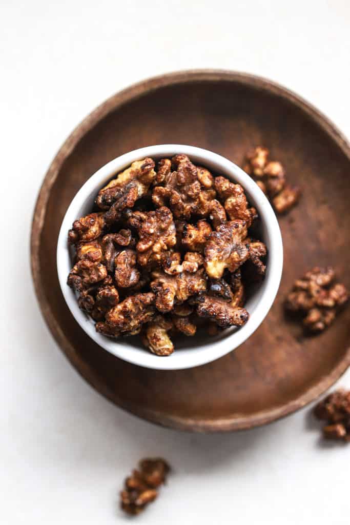 Maple cayenne toasted walnuts in small white bowl on small wooden plate with other walnuts sprinkled around