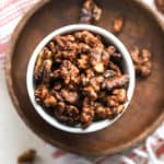 Maple cayenne toasted walnuts in small white bowl on small wooden plate with other walnuts sprinkled around, on white and pink linen