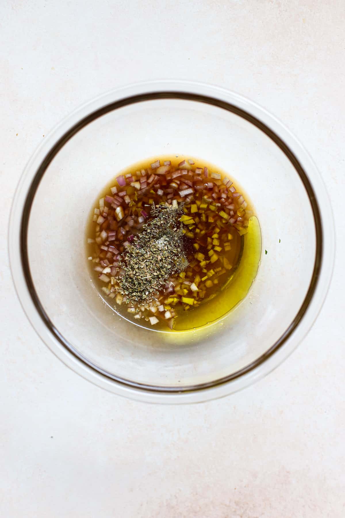 Extra virgin olive oil, sherry vinegar, lemon, minced onions, minced garlic, salt, pepper, and dried oregano in clear glass bowl on beige and white surface