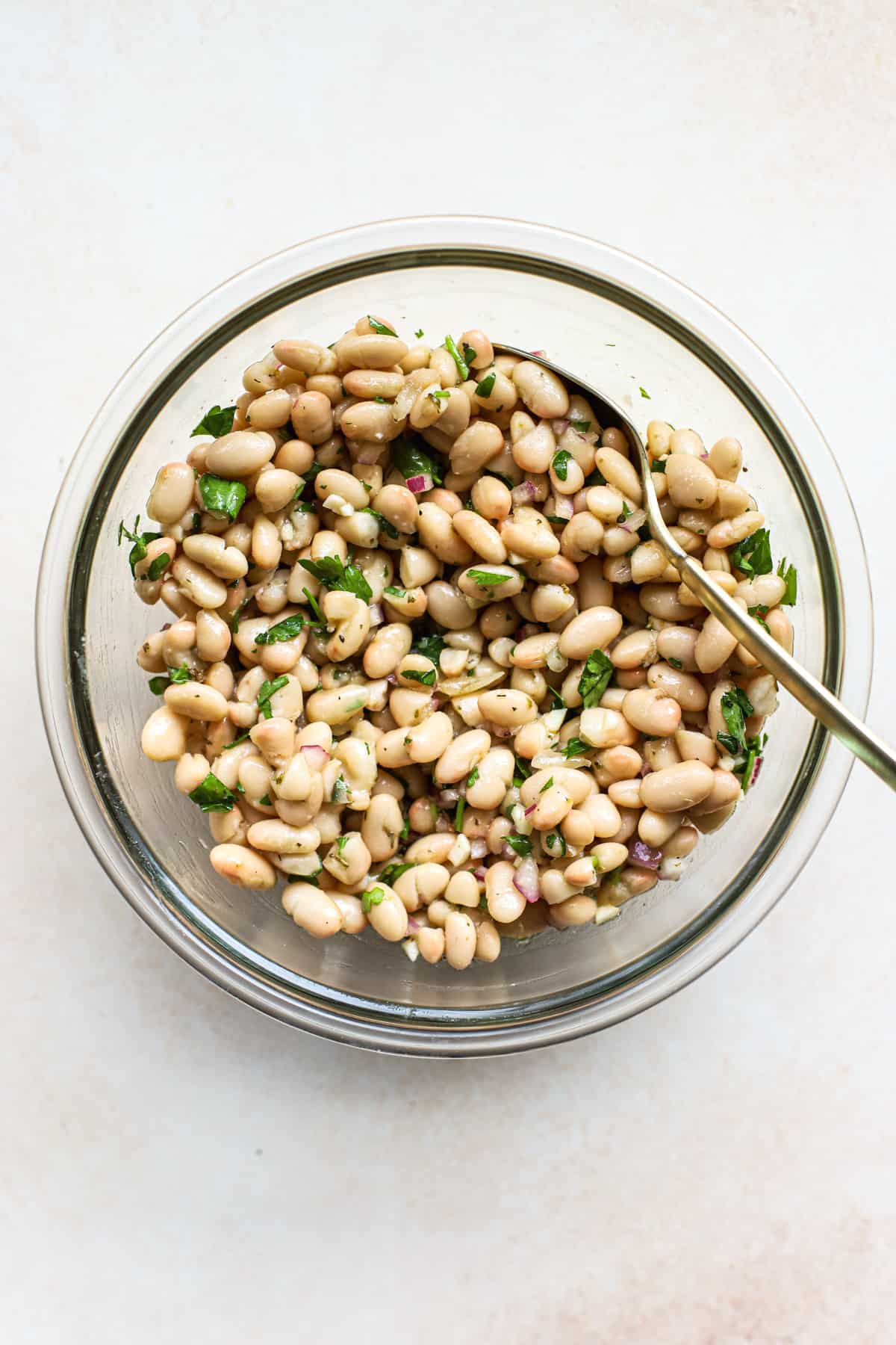 Marinated white beans stirred together with golden spoon in clear glass bowl, on beige and white surface
