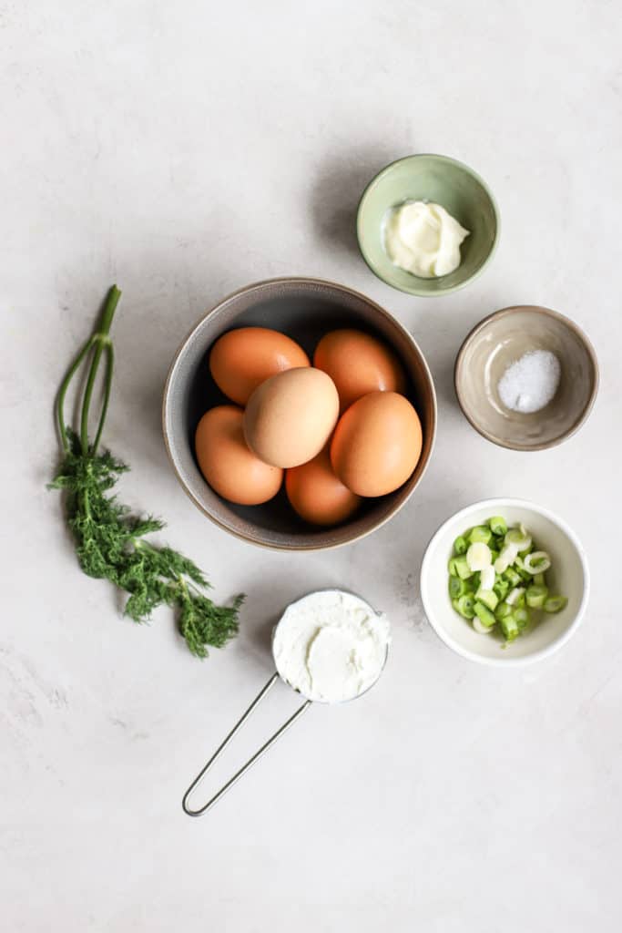 Eggs, fresh dill, Greek yogurt, mayo, salt, and green onions in pinch bowls on light gray and white surface