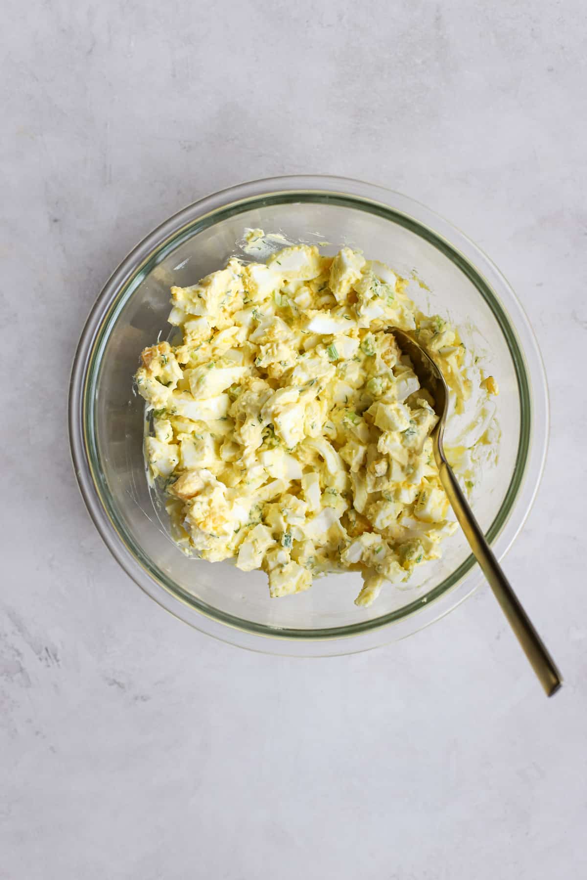 Healthy egg salad in clear glass bowl with spoon, on light gray surface