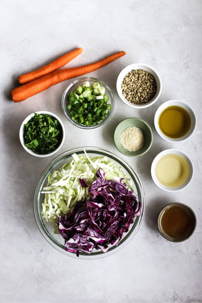 Healthy coleslaw ingredients (purple and green cabbage, carrots, green onions, parsley, sunflower seeds, sesame seeds, olive oil, rice vinegar, agave) on light gray surface