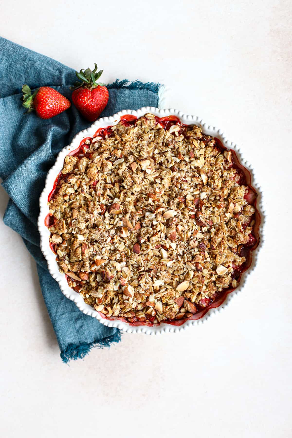Healthy strawberry crisp in cream-colored baking dish next to teal linen on beige and white surface with two fresh strawberries on the side