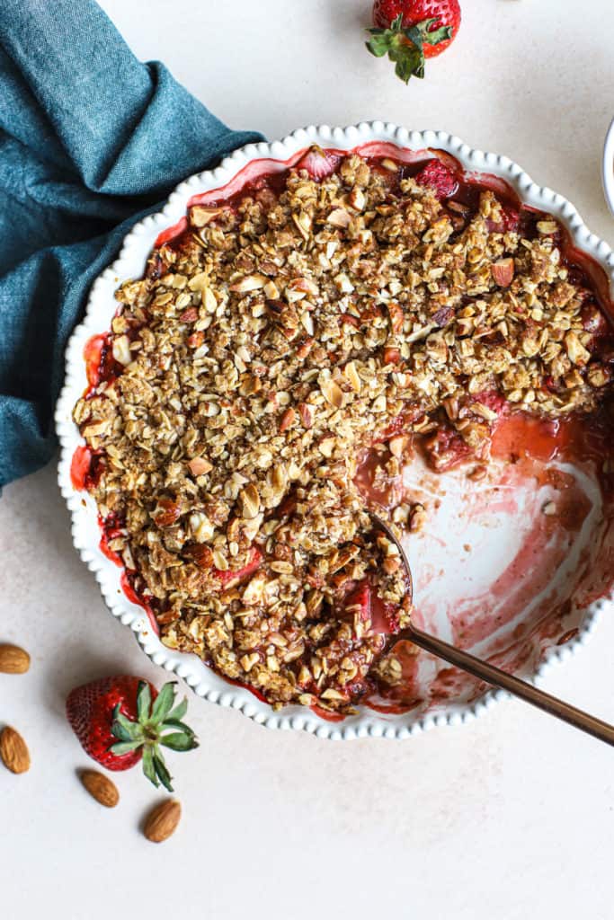 Healthy strawberry crisp in cream-colored pie dish large piece missing, copper serving spoon, and fresh strawberries and almonds on the side, next to teal linen