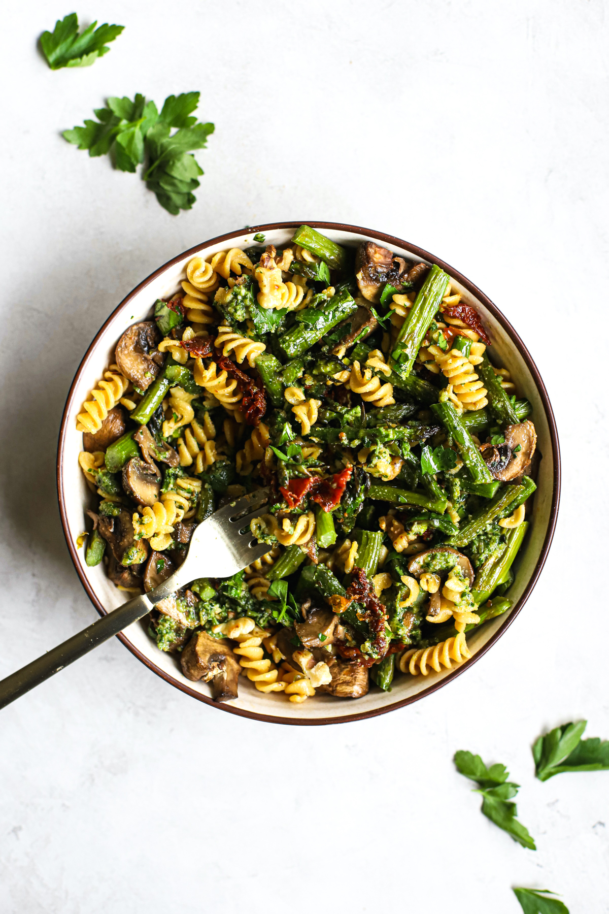 Pesto pasta with spring roasted veggies in beige bowl with golden fork on gray and white surface with parsley sprinkled around