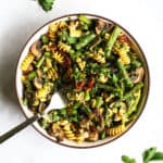 Pesto pasta with spring roasted veggies in beige bowl with golden fork on gray and white surface with parsley sprinkled around