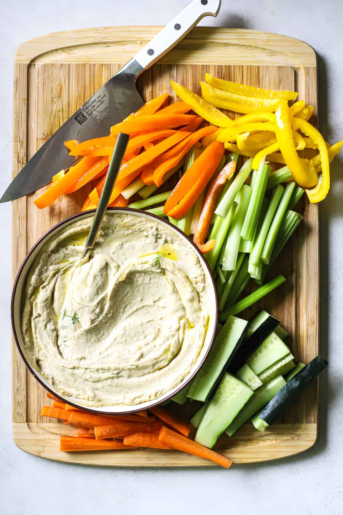 Carrots, cucumbers, celery, and bell peppers sliced on bamboo cutting board next to bowl with hummus and chef's knife