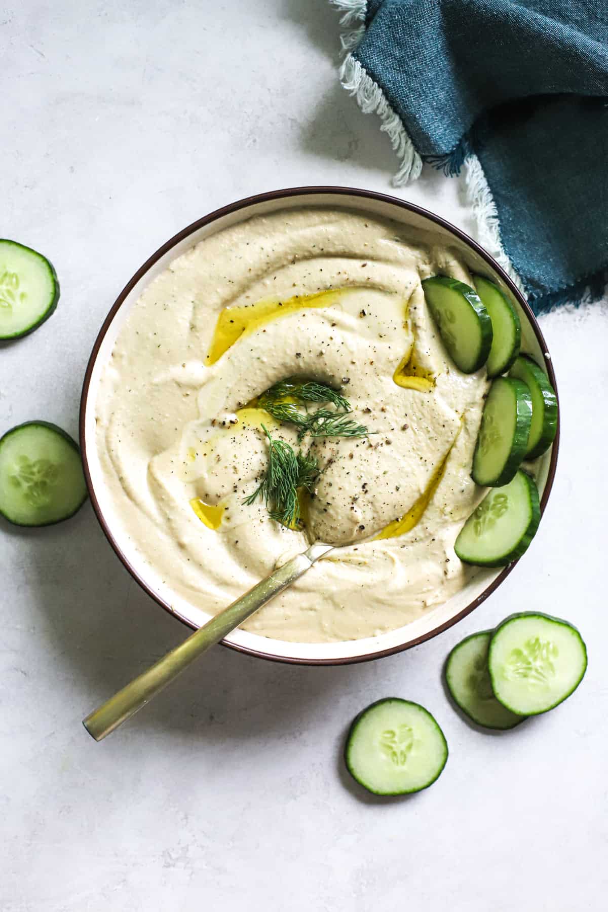 Lemon dill hummus in beige serving dish with olive oil drizzled on top, cucumbers dipped in and on the side, and golden spoon scooped into the hummus