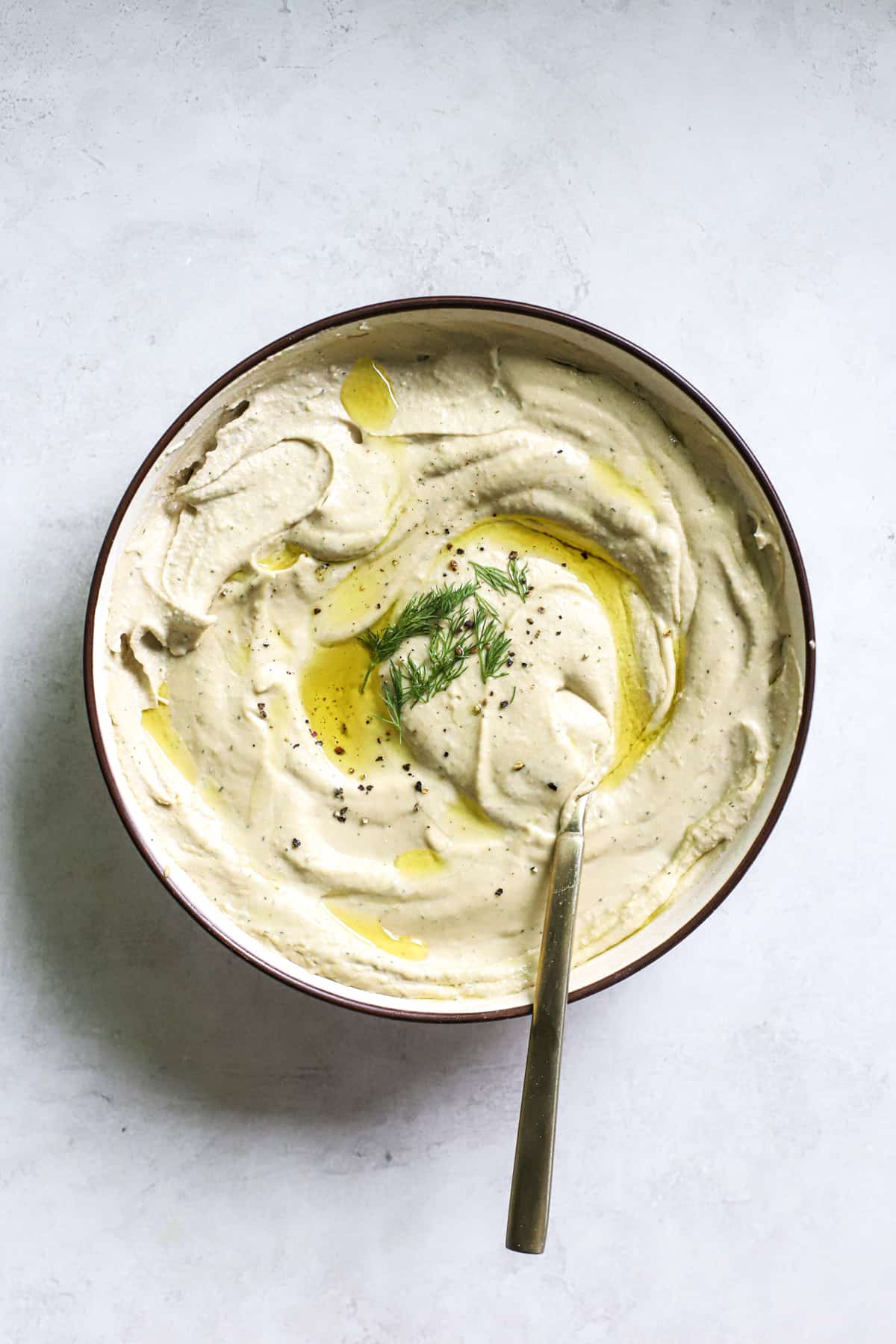 Lemon dill hummus in beige serving dish with olive oil drizzled on top, with golden spoon