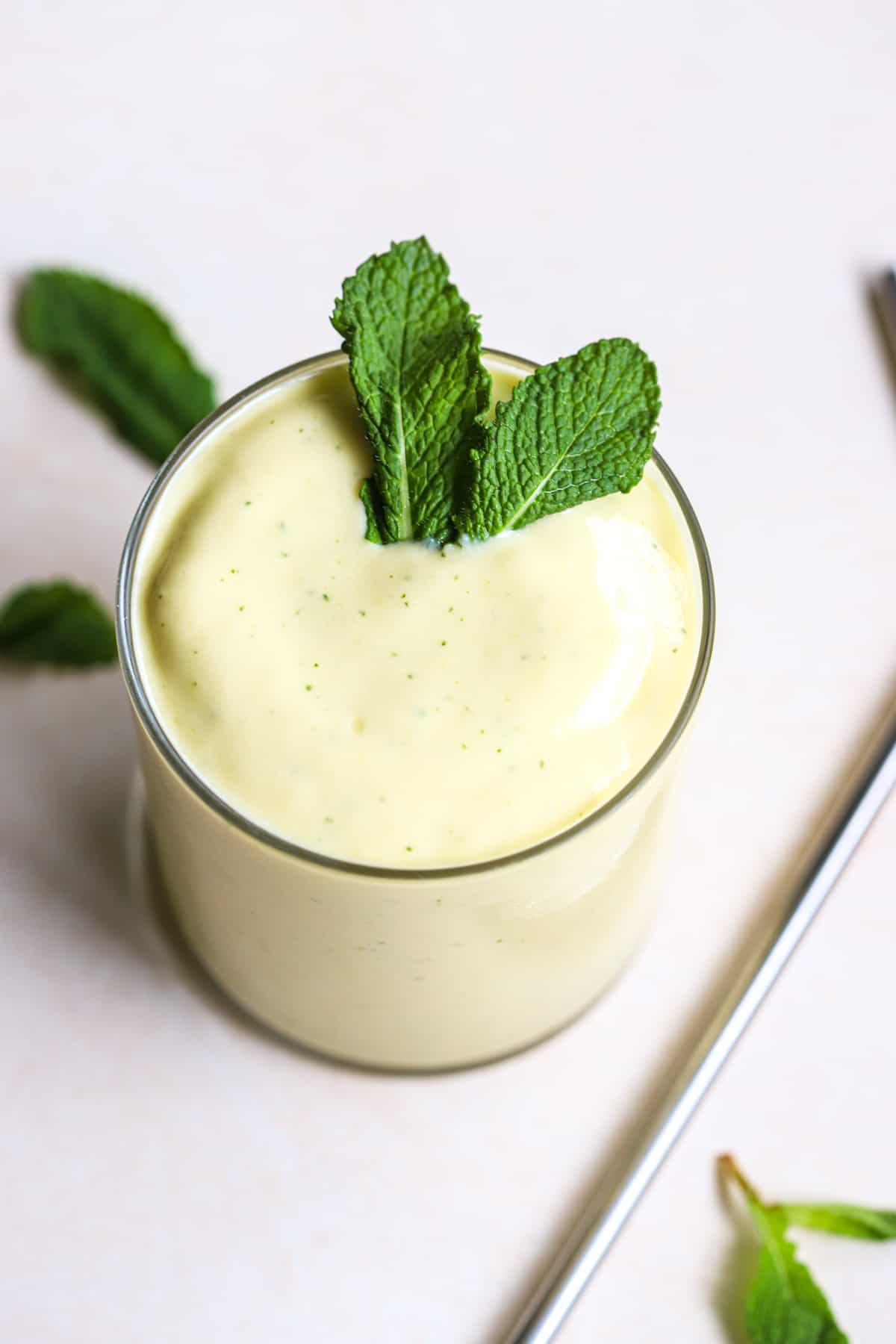 Mango mint smoothie in small glass with fresh mint leaves and stainless steel straw on side, on light beige and white surface