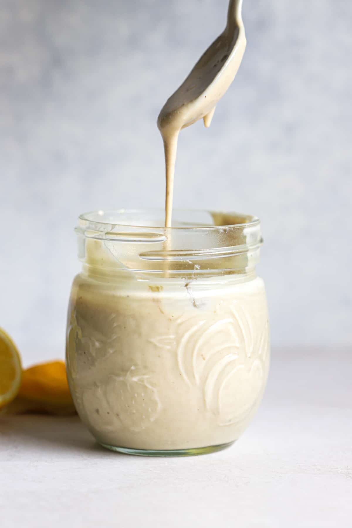 Tahini sauce dripping off of spoon into small glass jar, with lemons in the background