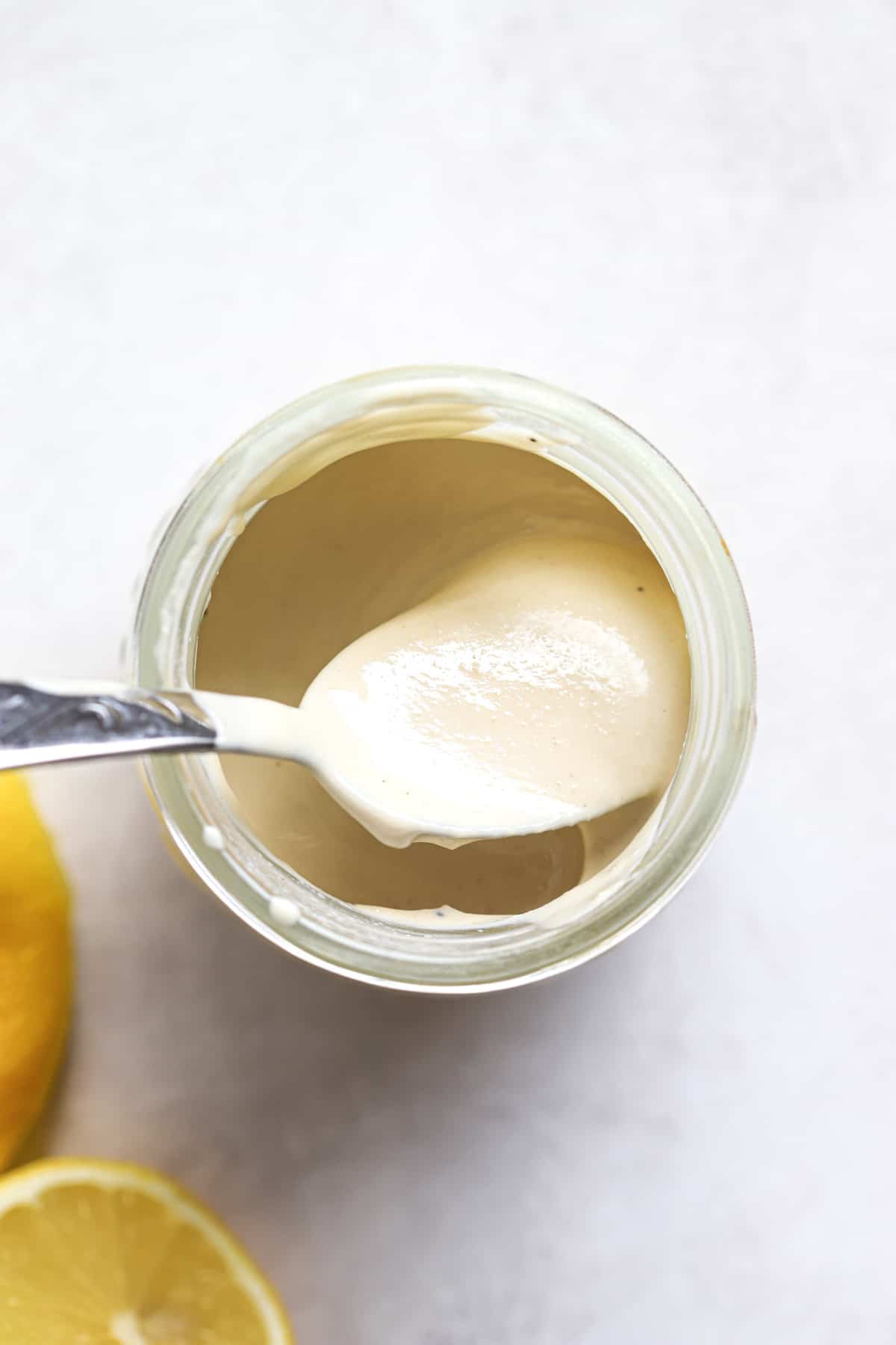 Tahini sauce in small glass jar with spoon and lemons on the side, on gray and white surface