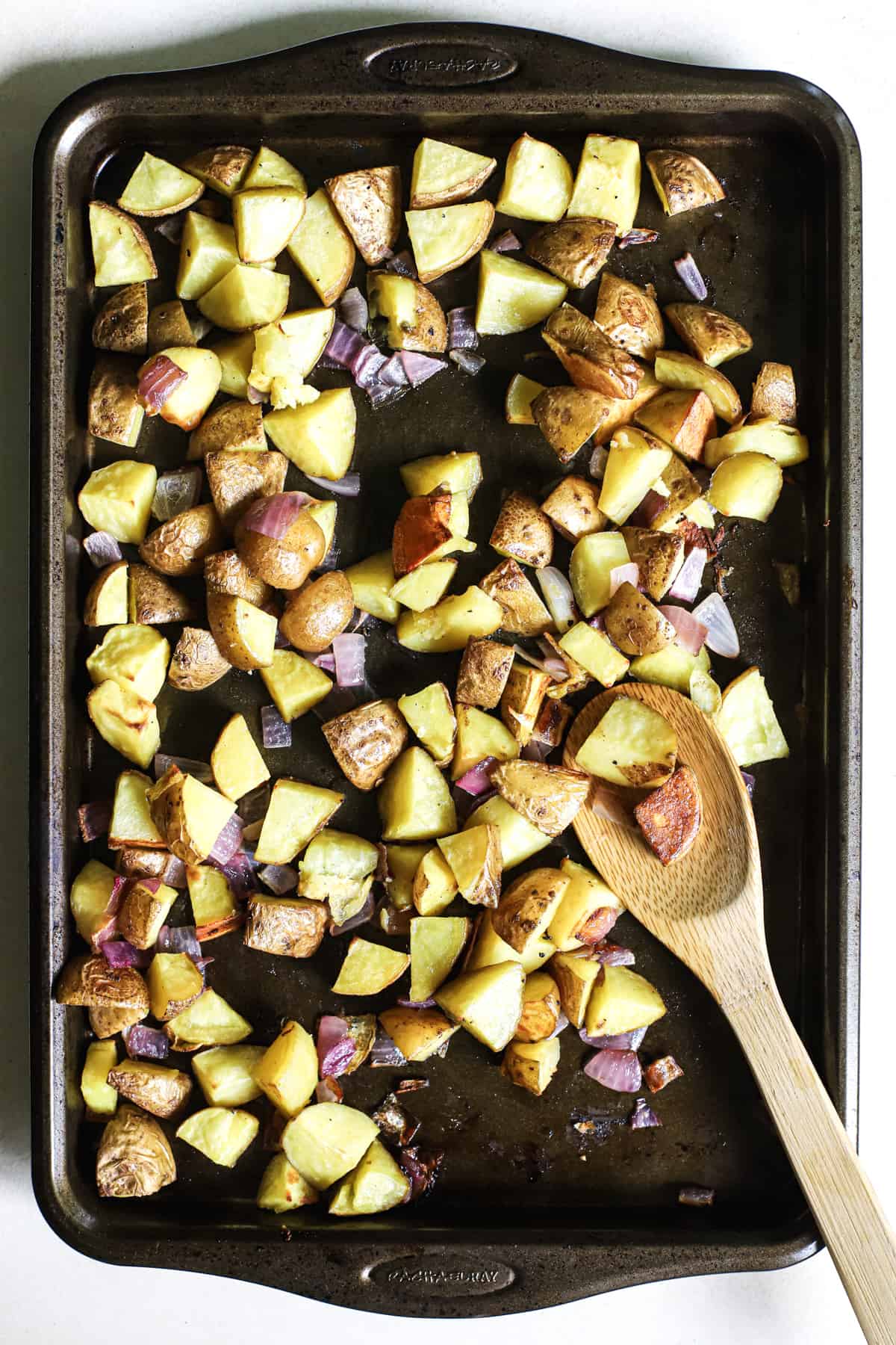 Roasted potatoes and red onions on baking sheet with wooden spoon scooping some potatoes