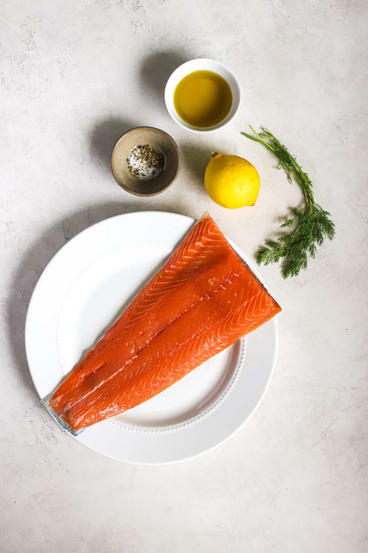 Fresh salmon on white plate, with lemon, fresh dill, salt, pepper, and olive oil on side