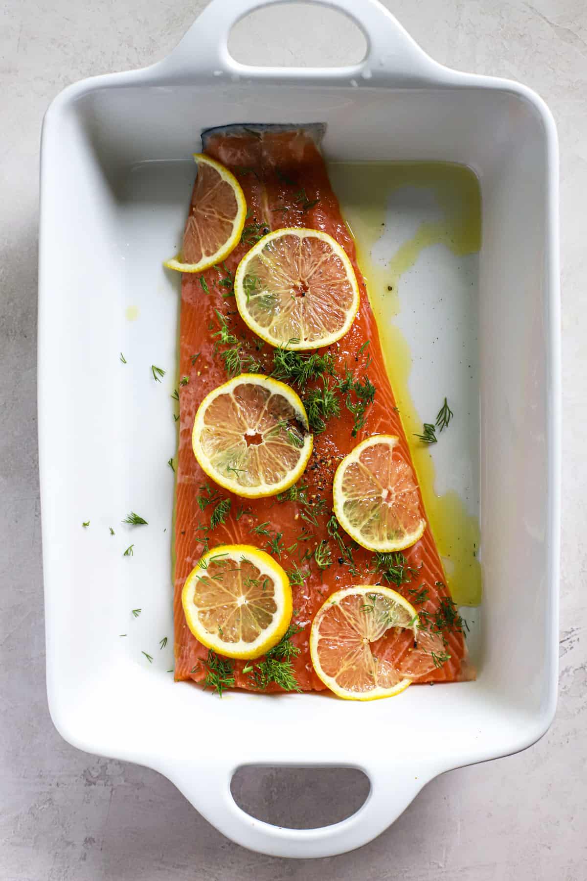 Fresh salmon with olive oil, salt, pepper, fresh dill, and thinly sliced lemon in white ceramic baking dish