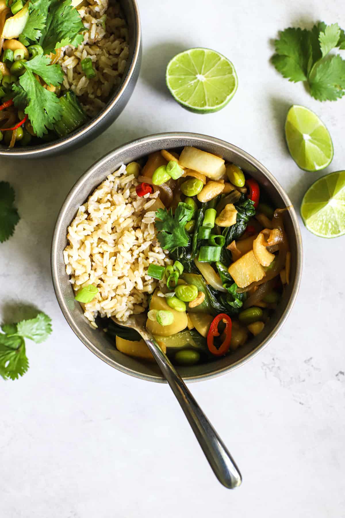 Two gray bowls with pad pak, served with brown rice, with limes wedges and cilantro on the side