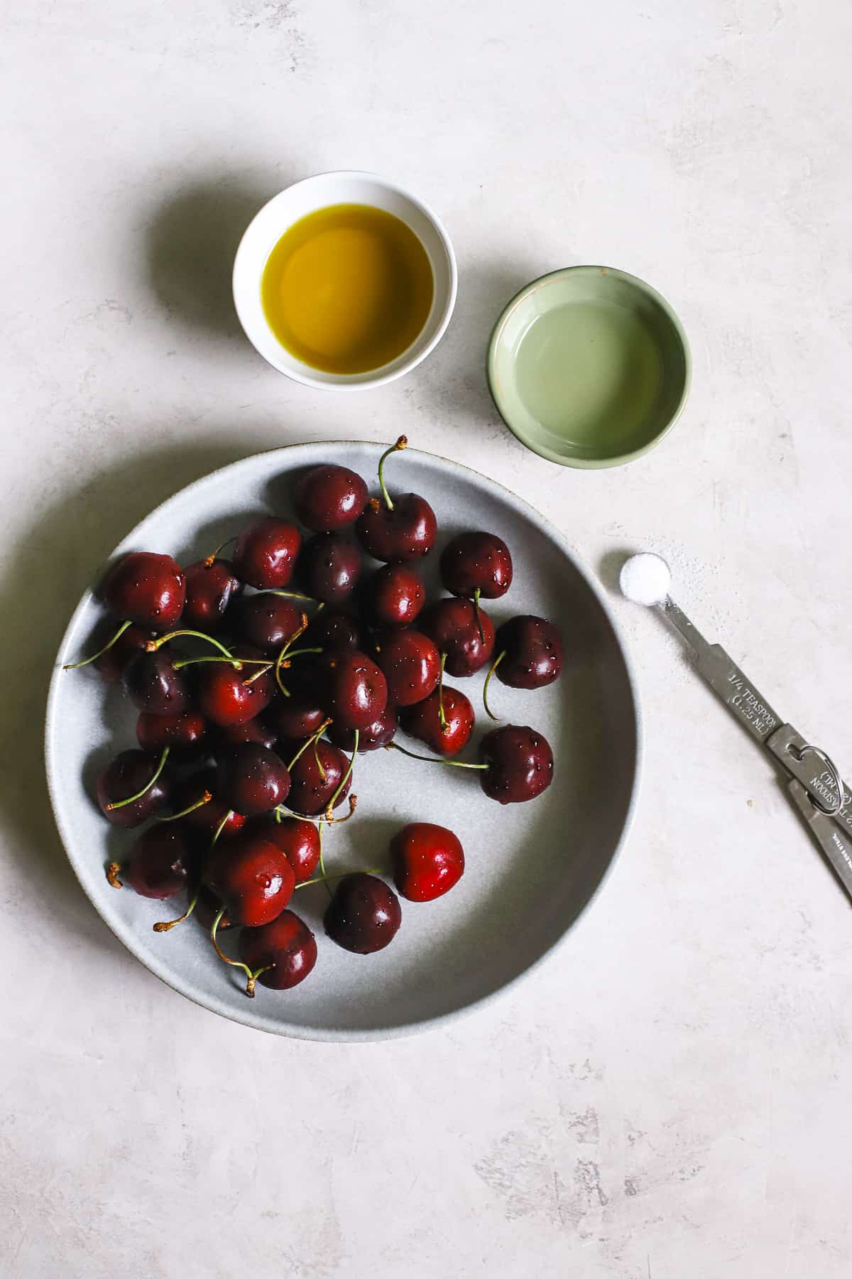 Vinaigrette ingredients: sweet cherries, olive oil, light vinegar, and salt on gray-white surface