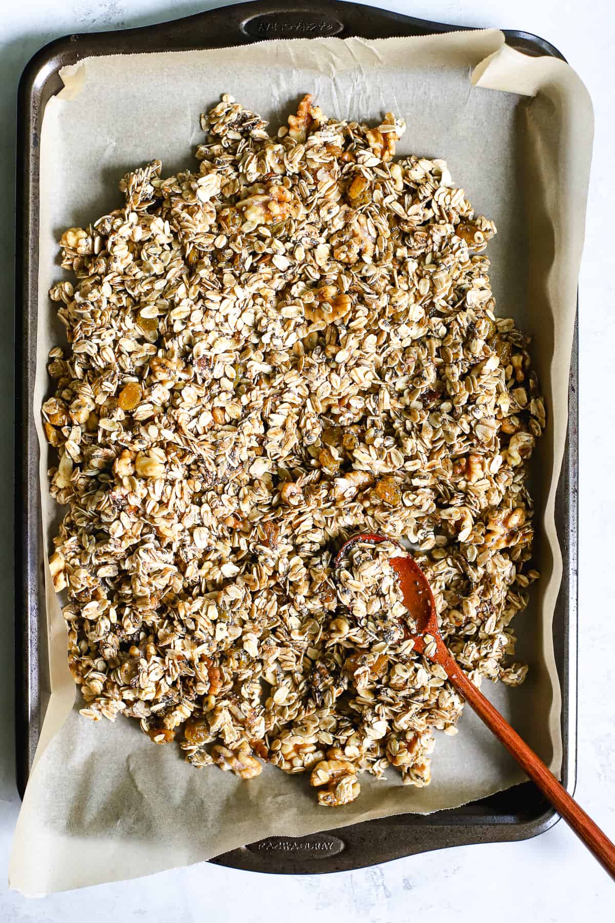 Maple cinnamon walnut granola spreading onto parchment-lined sheet pan with wooden spoon