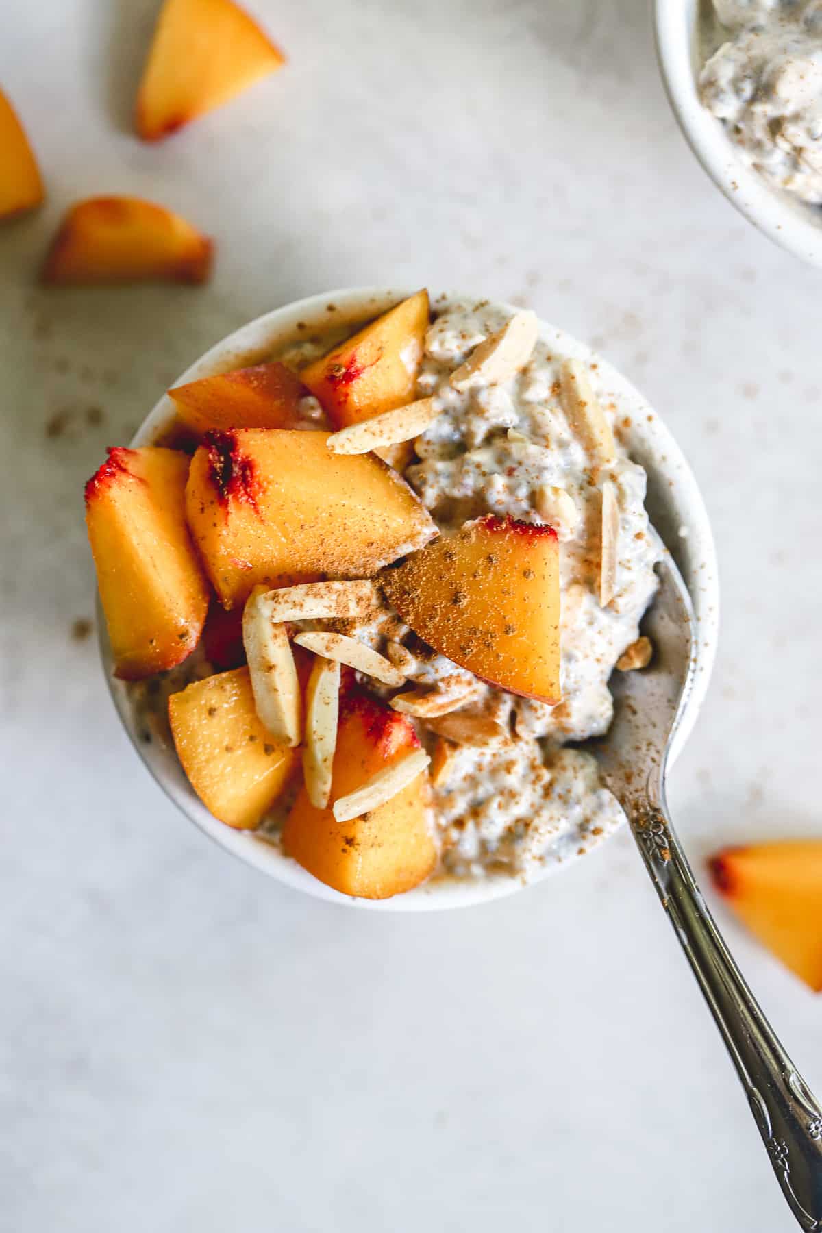 Peach pie overnight oats in small white bowl with diced fresh peaches, slivered almonds, and a dusting of cinnamon