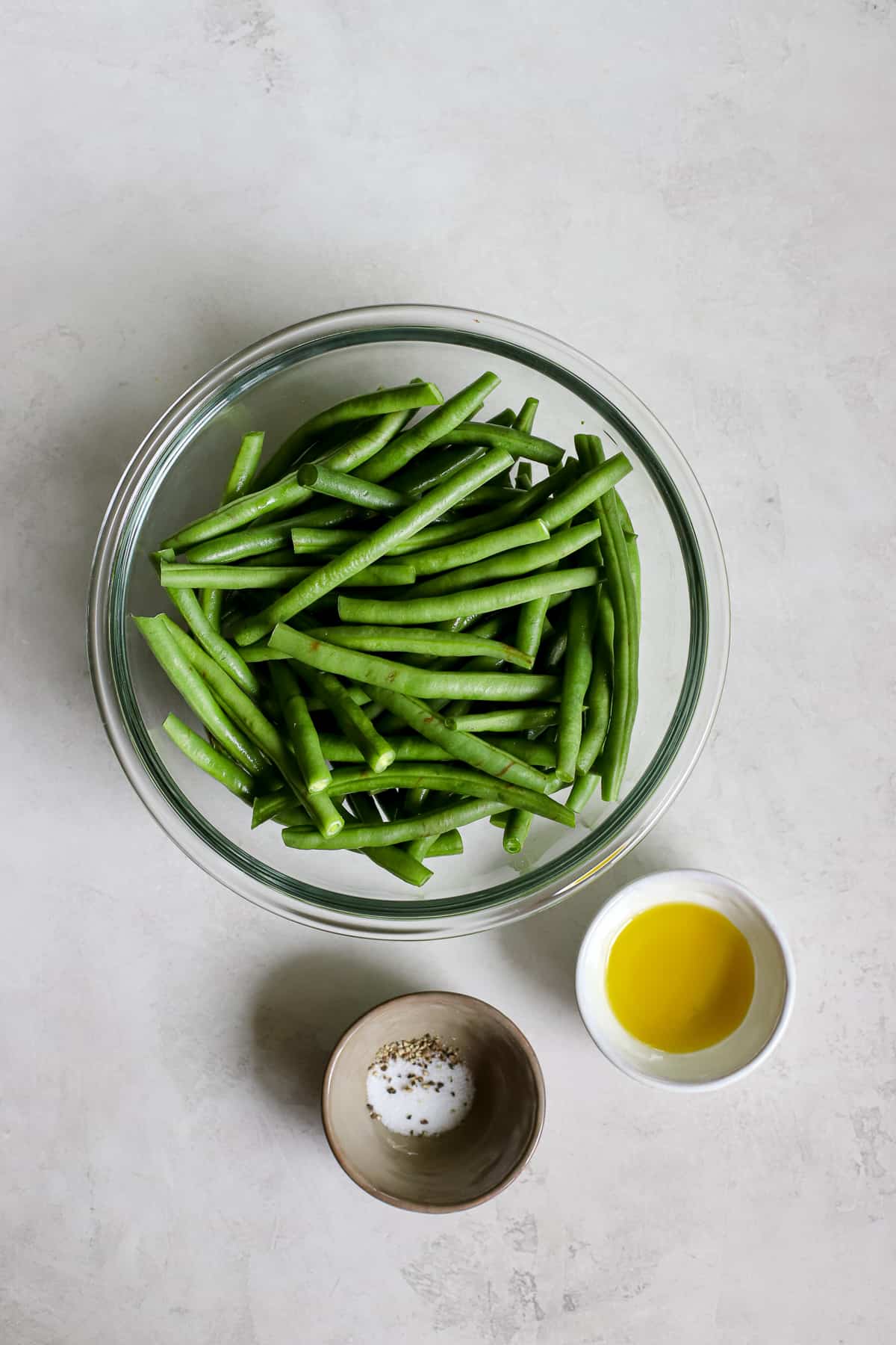 Fresh green beans, salt, pepper, and olive oil in bowls on gray and white surfact